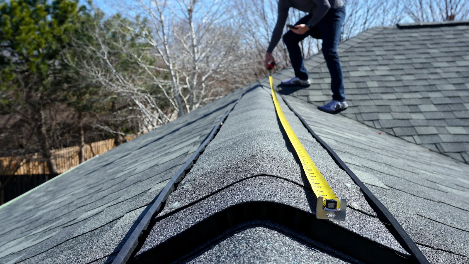 Illustrative roofer measuring along a gray shingle roof ridge with a yellow tape measure